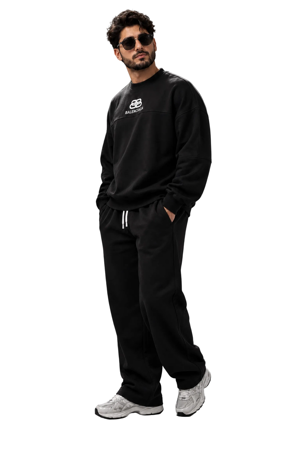 A man wearing a black crewneck sweatshirt with a small white chest logo, black sweatpants with white drawstrings, and white sneakers, standing against a dark gray studio background.