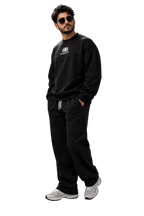 A man wearing a black crewneck sweatshirt with a small white chest logo, black sweatpants with white drawstrings, and white sneakers, standing against a dark gray studio background.