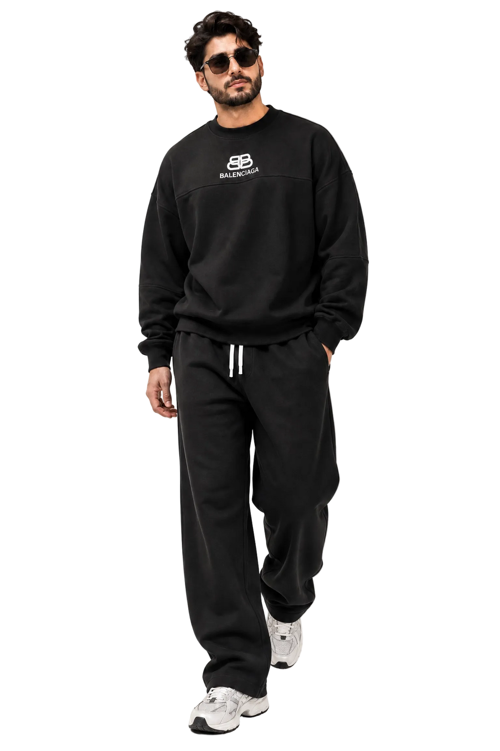 A man modeling a black Balenciaga sweatshirt and matching sweatpants set, featuring a small white logo on the chest and white drawstrings on the pants, set against a minimalist studio background with diagonal shadows.