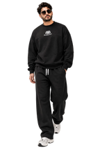 A man modeling a black Balenciaga sweatshirt and matching sweatpants set, featuring a small white logo on the chest and white drawstrings on the pants, set against a minimalist studio background with diagonal shadows.