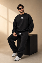 A man wearing a black Balenciaga sweatshirt and matching sweatpants sits on a black block against a beige studio backdrop. The Balenciaga logo is visible on the chest of the sweatshirt, and he is wearing white sneakers.