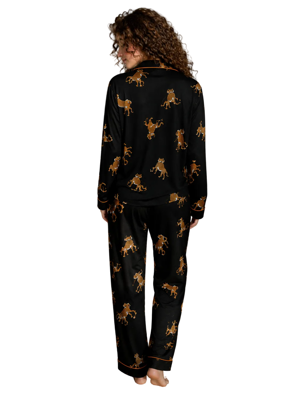 A woman stands barefoot in a bedroom wearing a black pajama set with an allover gold and brown leopard print. The photo is taken from behind, showing the back of the long-sleeve top and matching pants.