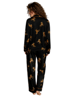 A woman stands barefoot in a bedroom wearing a black pajama set with an allover gold and brown leopard print. The photo is taken from behind, showing the back of the long-sleeve top and matching pants.