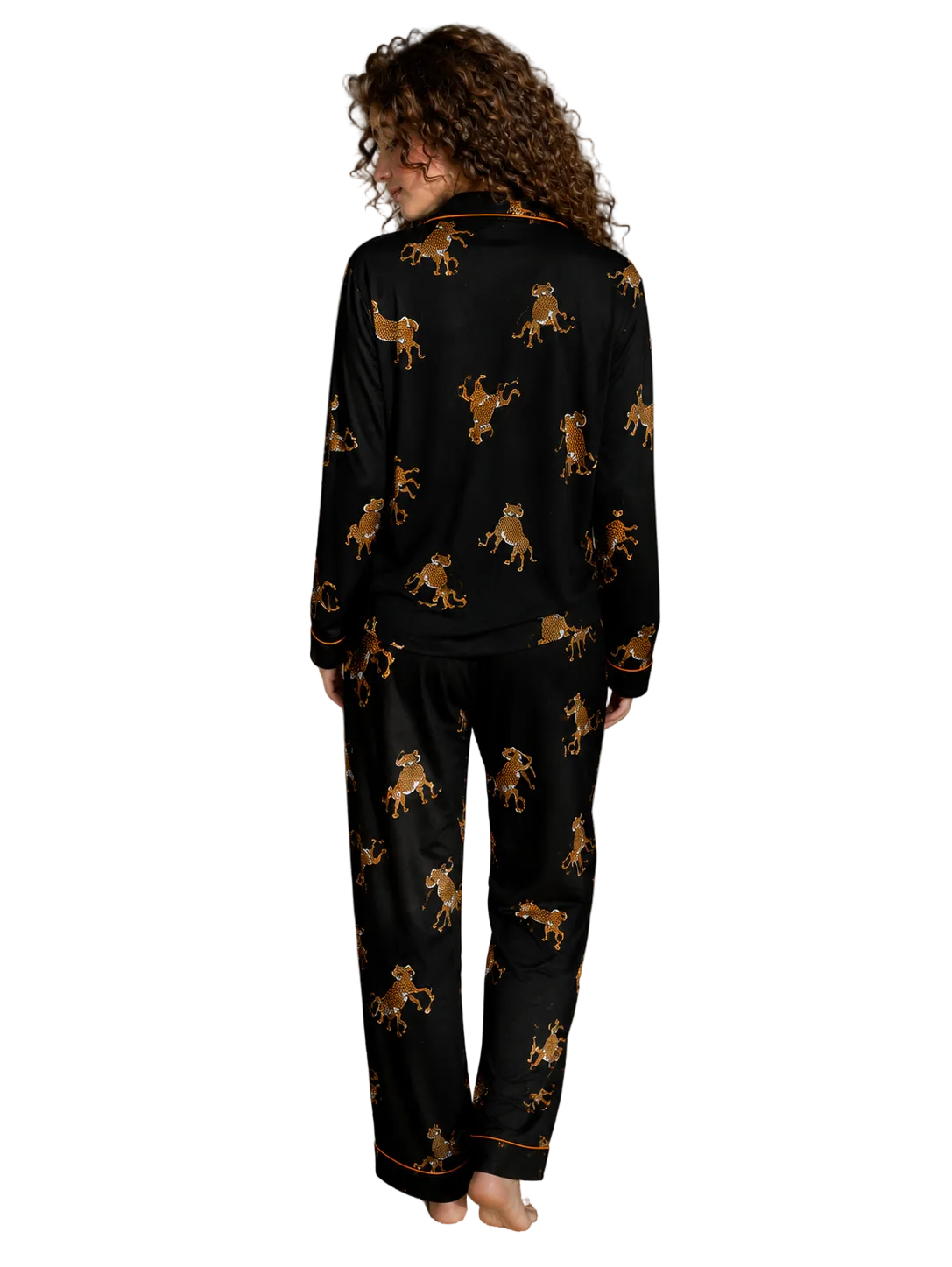A woman stands barefoot in a bedroom wearing a black pajama set with an allover gold and brown leopard print. The photo is taken from behind, showing the back of the long-sleeve top and matching pants.