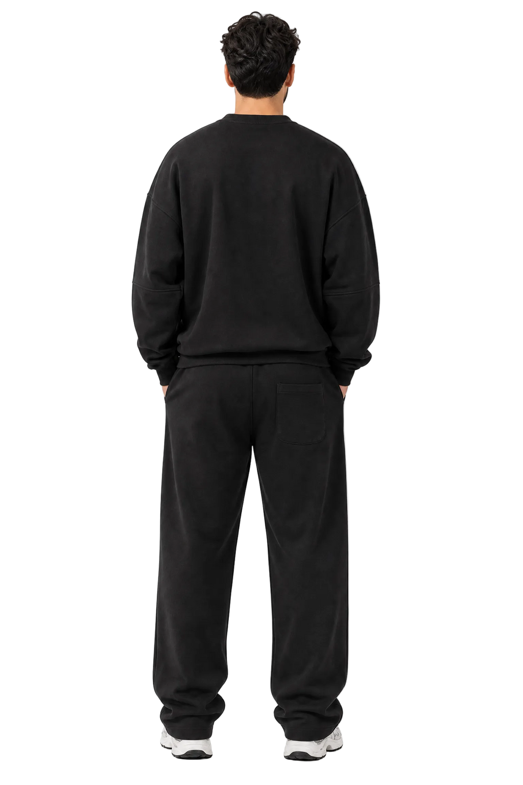 A man viewed from behind wearing a black crewneck sweatshirt and matching black sweatpants, standing against a plain light gray studio background.