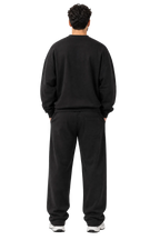 A man viewed from behind wearing a black crewneck sweatshirt and matching black sweatpants, standing against a plain light gray studio background.