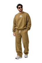 A man modeling a matching olive green sweatshirt and jogger pants set with white drawstrings and a white chest logo, styled with gray sneakers against a gray studio backdrop.