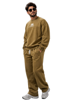 A man wearing a matching olive-brown sweatshirt and sweatpants set with white sneakers, standing against a dark gray studio background.
