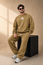 A man wearing a matching olive-brown sweatshirt and jogger pants set with white drawstrings and a white 'SB Balenciaga' logo on the chest, sitting on a black block against a beige studio backdrop.