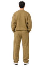A man viewed from behind wearing a matching two-piece lounge set consisting of a crewneck sweatshirt and relaxed-fit pants in a warm khaki-brown color, set against a light gray studio background.