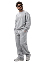 A man wearing a light gray crewneck sweatshirt and matching sweatpants with white sneakers, standing against a dark gray studio background.