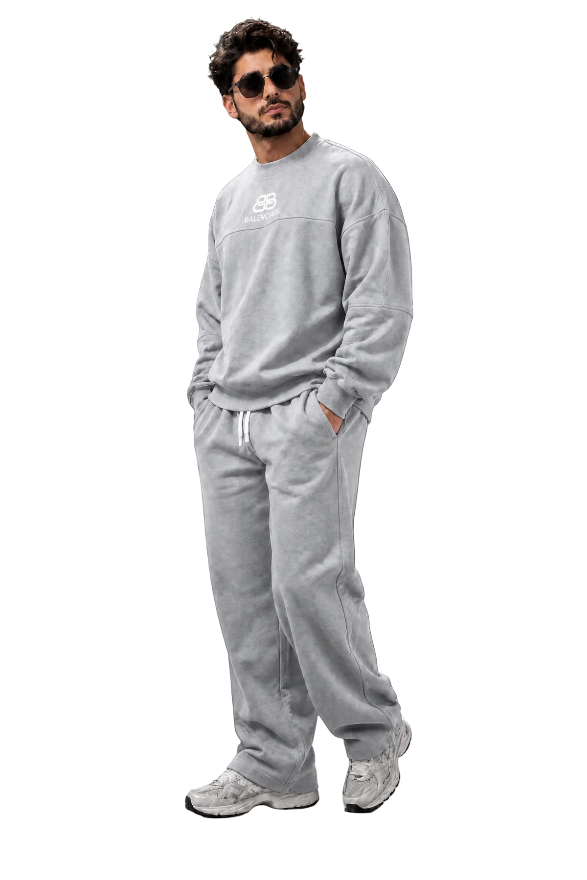 A man wearing a light gray crewneck sweatshirt and matching sweatpants with white sneakers, standing against a dark gray studio background.