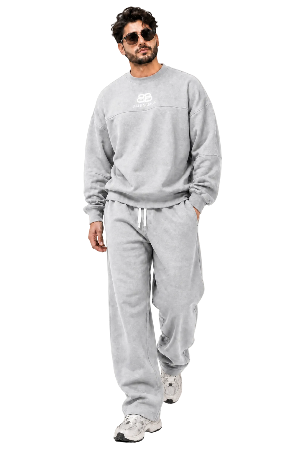 A man wearing a light gray sweatshirt and matching sweatpants with white sneakers, walking against a neutral beige background with geometric shadows.