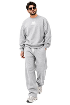 A man wearing a light gray sweatshirt and matching sweatpants with white sneakers, walking against a neutral beige background with geometric shadows.