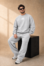 A man wearing a light gray crewneck sweatshirt and matching sweatpants sits on a black block against a textured beige studio backdrop. The sweatshirt features a white 'BB' logo on the chest, and the model is wearing white sneakers.