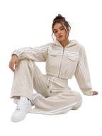 A woman sits on a light gray studio floor wearing a beige hooded tracksuit with white stripes and white sneakers. The tracksuit has a zip front, chest pockets, and matching wide-leg pants with white side stripes.