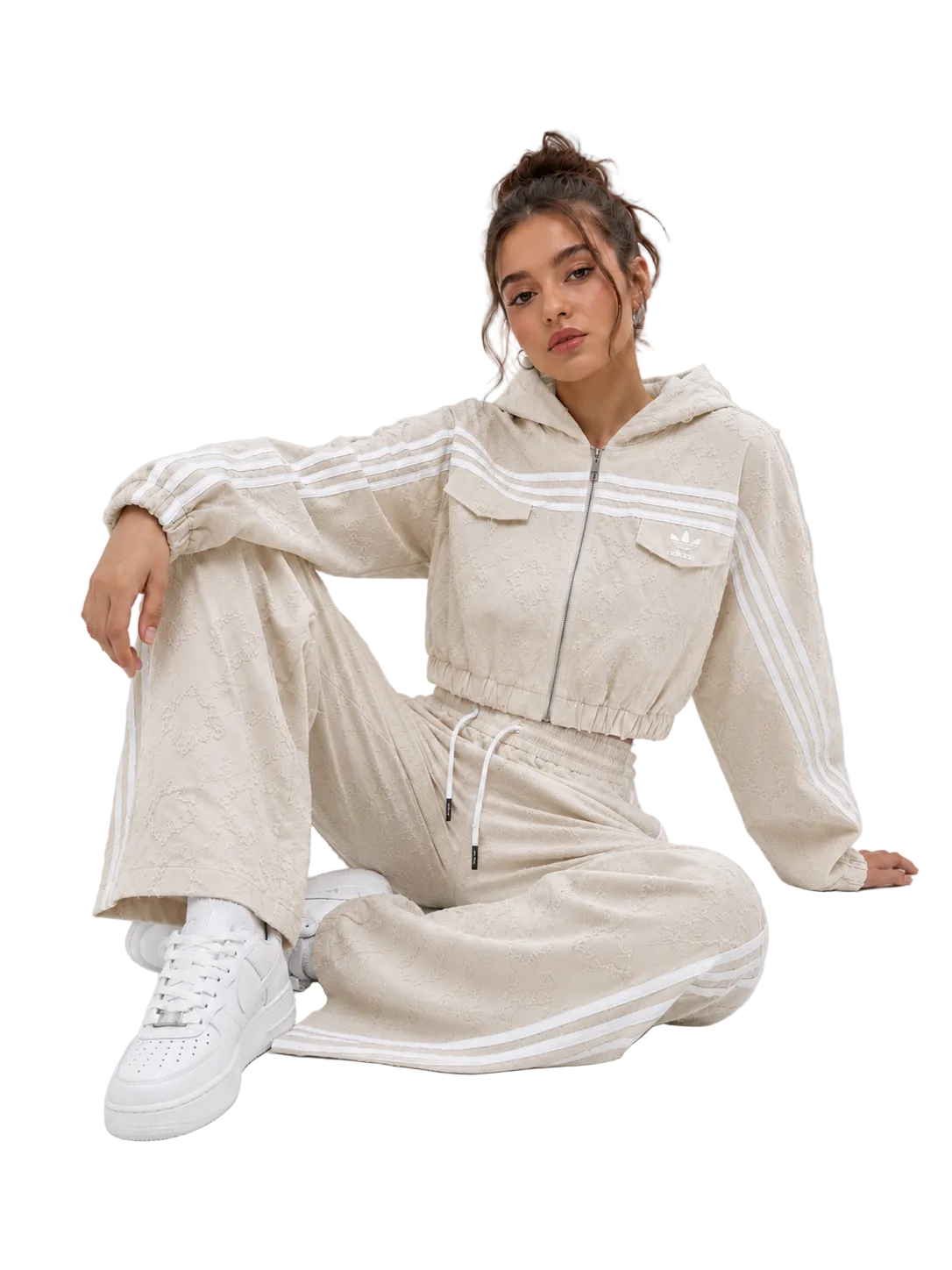 A woman sits on a light gray studio floor wearing a beige hooded tracksuit with white stripes and white sneakers. The tracksuit has a zip front, chest pockets, and matching wide-leg pants with white side stripes.