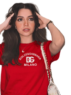 A woman wearing a red Dolce & Gabbana Milano t-shirt with a white logo print, posing with a patterned shoulder bag against a modern beige wall.