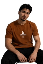 A man wearing a rust-brown short-sleeve t-shirt with a white graphic print on the chest, sitting in a modern room with a gray wall and a black leather sofa in the background.