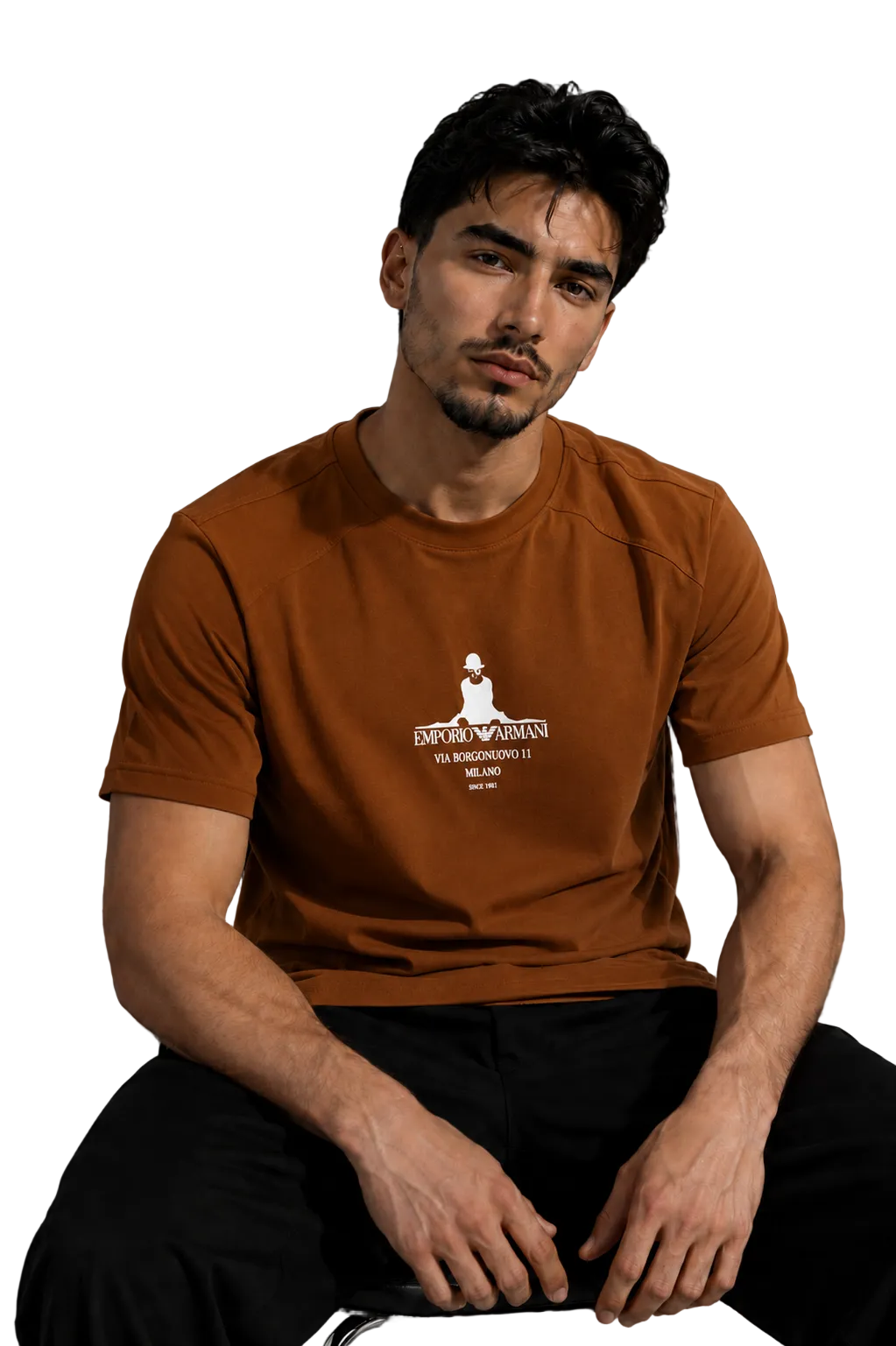 A man wearing a rust-brown short-sleeve t-shirt with a white graphic print on the chest, sitting in a modern room with a gray wall and a black leather sofa in the background.