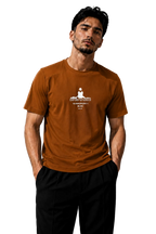 A man wearing a rust-orange crewneck t-shirt with a white graphic print of a seated figure and Sanskrit text, paired with black pants, standing against a textured gray wall with dramatic side lighting.