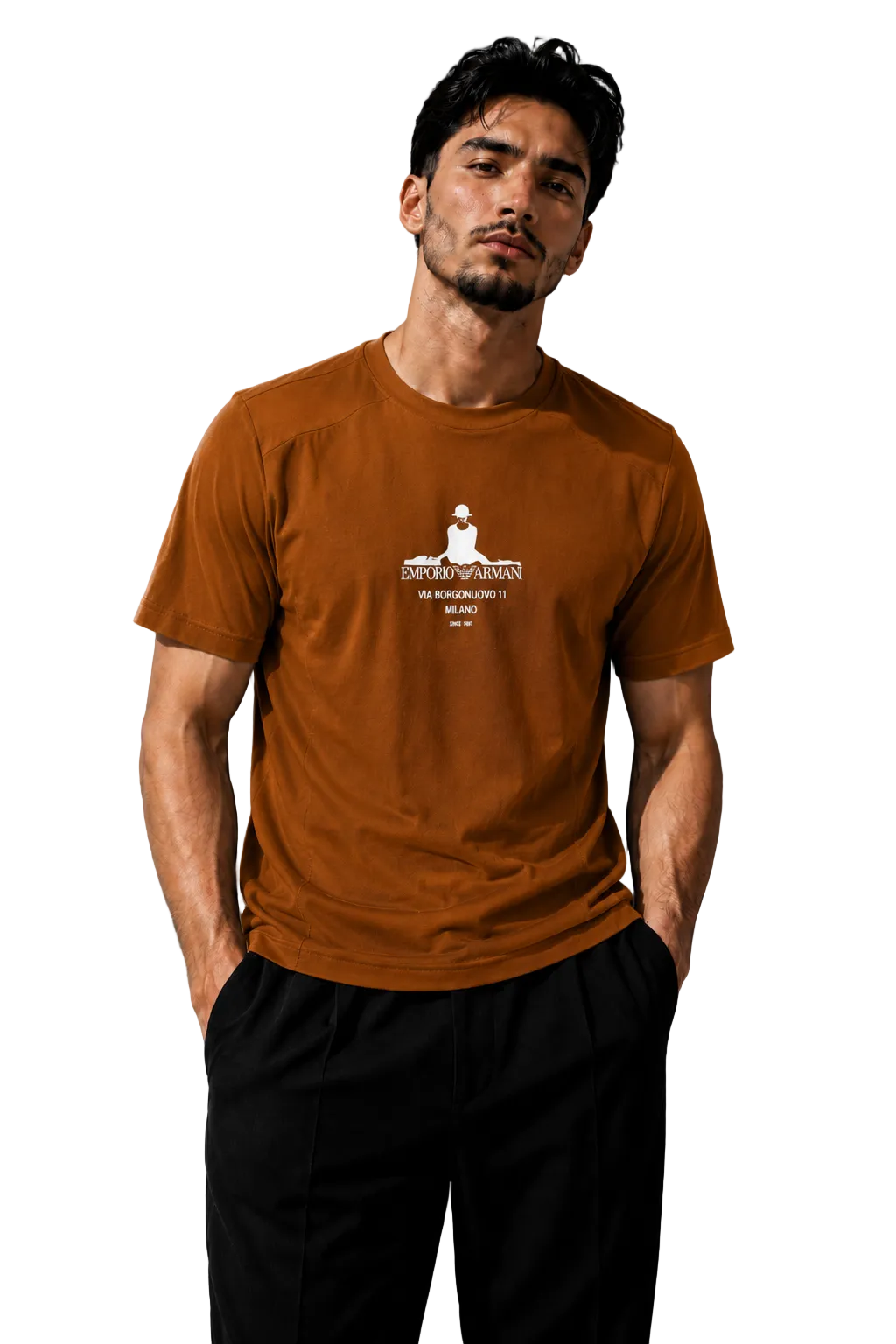 A man wearing a rust-orange crewneck t-shirt with a white graphic print of a seated figure and Sanskrit text, paired with black pants, standing against a textured gray wall with dramatic side lighting.