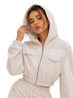A woman wearing a beige textured zip-up hooded jacket with white stripes on the sleeves and chest, paired with matching beige joggers, against a neutral gray background.