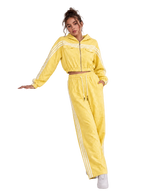 A woman models a bright yellow two-piece tracksuit consisting of a cropped zip-up hoodie with white stripes and matching wide-leg sweatpants with white side stripes, posed next to a white display block in a studio setting.