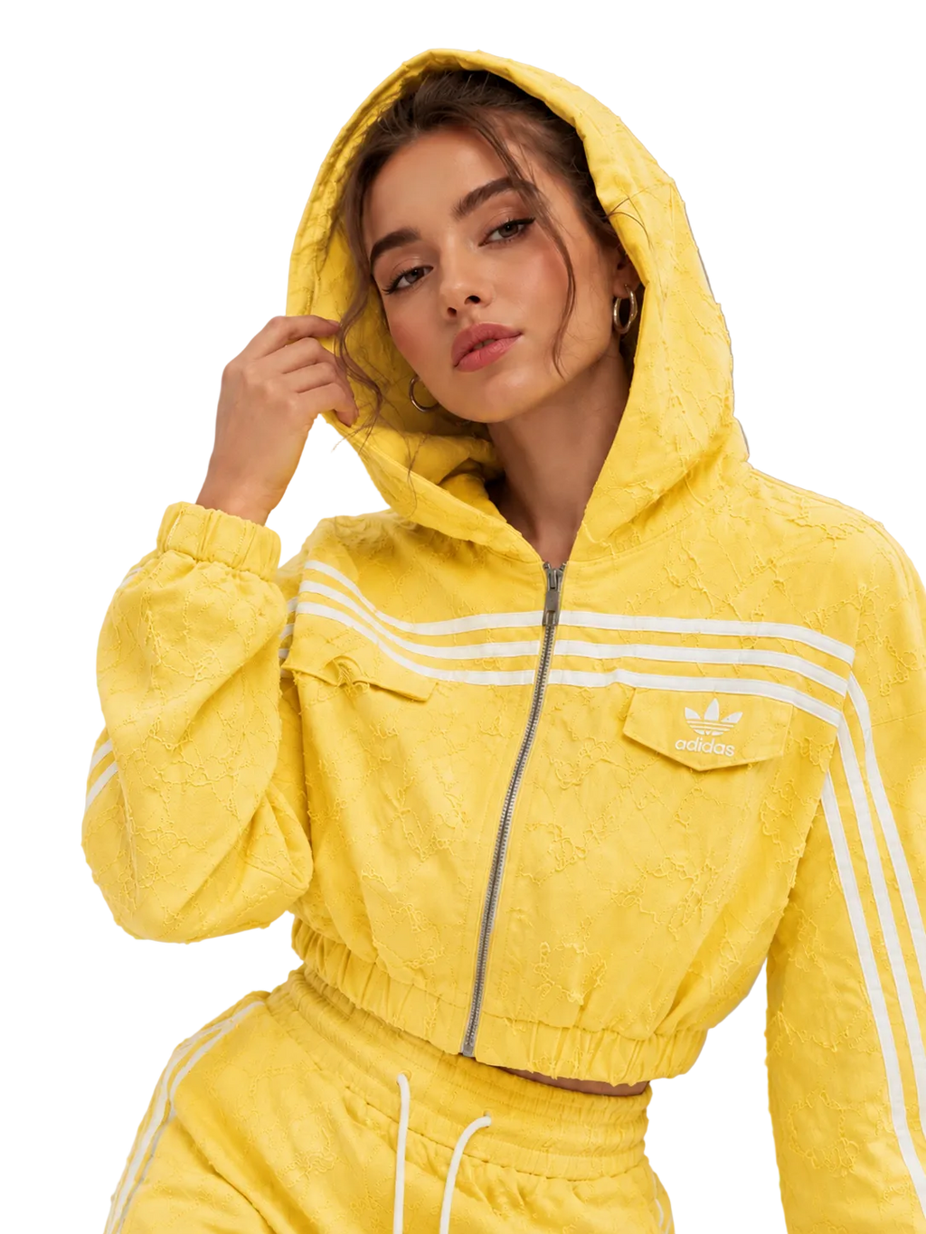 A woman wearing a bright yellow textured Adidas hooded track jacket with white stripes and matching yellow track pants, posed against a neutral gray studio background.
