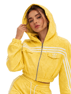 A woman wearing a bright yellow textured Adidas hooded track jacket with white stripes and matching yellow track pants, posed against a neutral gray studio background.