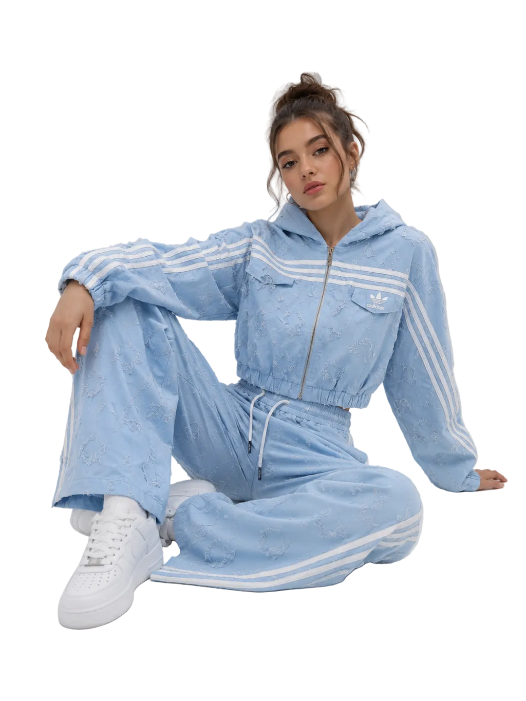 A woman sits on a light gray studio floor wearing a light blue embroidered tracksuit with white stripes, featuring a cropped zip-up hoodie and matching wide-leg pants, paired with white sneakers.