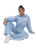 A woman sits on a light gray studio floor wearing a light blue embroidered tracksuit with white stripes, featuring a cropped zip-up hoodie and matching wide-leg pants, paired with white sneakers.