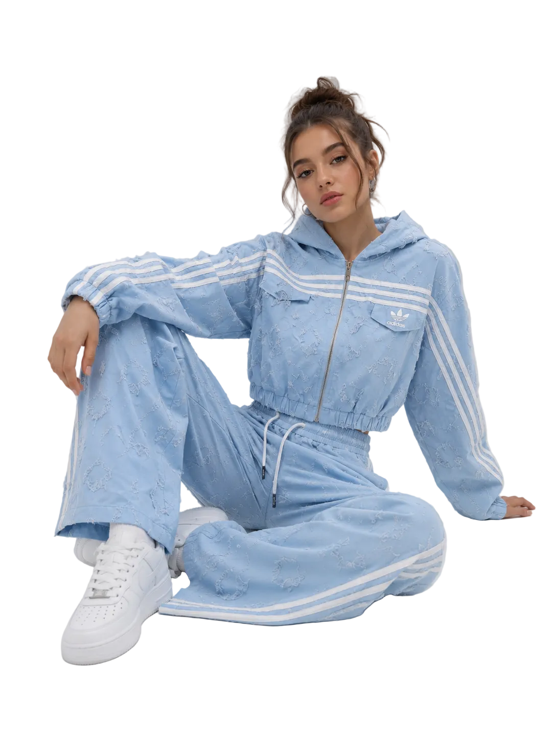A woman sits on a light gray studio floor wearing a light blue embroidered tracksuit with white stripes, featuring a cropped zip-up hoodie and matching wide-leg pants, paired with white sneakers.