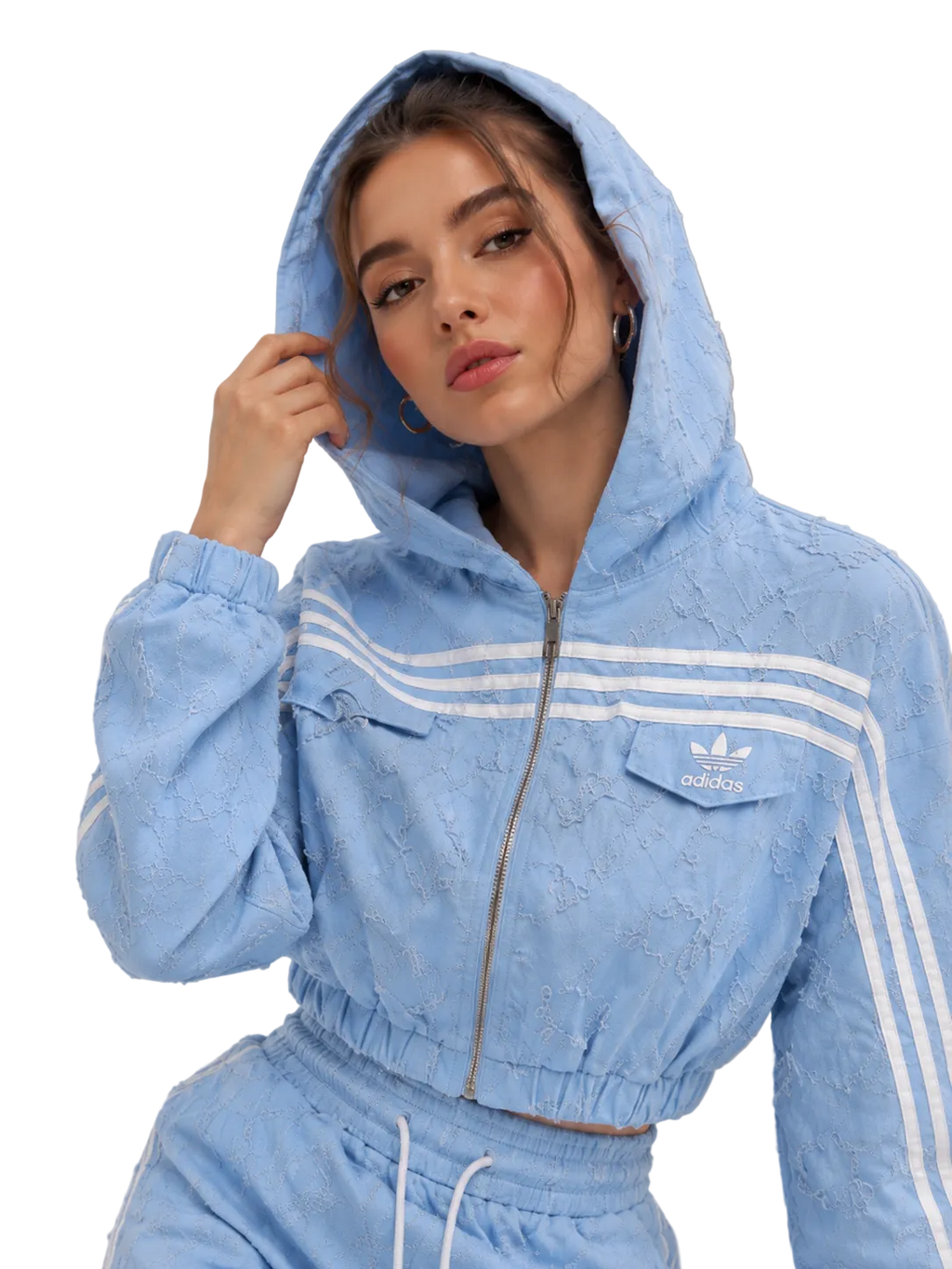 A woman wearing a light blue textured Adidas tracksuit with white stripes, featuring a cropped zip-up hoodie and matching jogger pants, set against a neutral studio background.