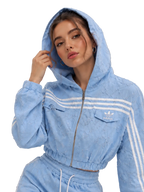 A woman wearing a light blue textured Adidas tracksuit with white stripes, featuring a cropped zip-up hoodie and matching jogger pants, set against a neutral studio background.