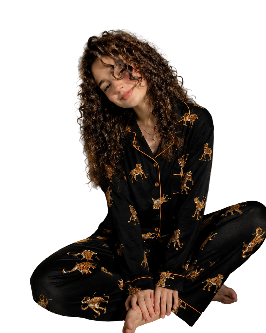 A woman with long curly brown hair sits cross-legged on a white carpet in a bedroom, wearing a black pajama set with a gold and brown leopard print and gold piping. The background shows a bed with white sheets and a bedside table.