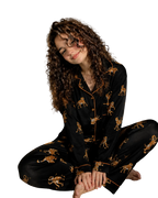 A woman with long curly brown hair sits cross-legged on a white carpet in a bedroom, wearing a black pajama set with a gold and brown leopard print and gold piping. The background shows a bed with white sheets and a bedside table.