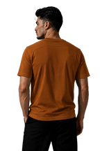 A man wearing a rust-orange short-sleeve t-shirt and black pants, shown from the back against a textured gray studio background.