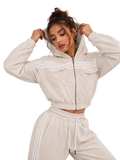 A woman modeling a light beige cropped zip-up hoodie with white stripes and matching jogger pants against a neutral gray studio background.