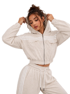 A woman modeling a light beige cropped zip-up hoodie with white stripes and matching jogger pants against a neutral gray studio background.