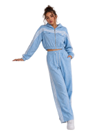 A woman models a light blue two-piece tracksuit featuring a cropped zip-up hoodie with white stripes and matching wide-leg sweatpants with white side stripes, posing next to a white display block.