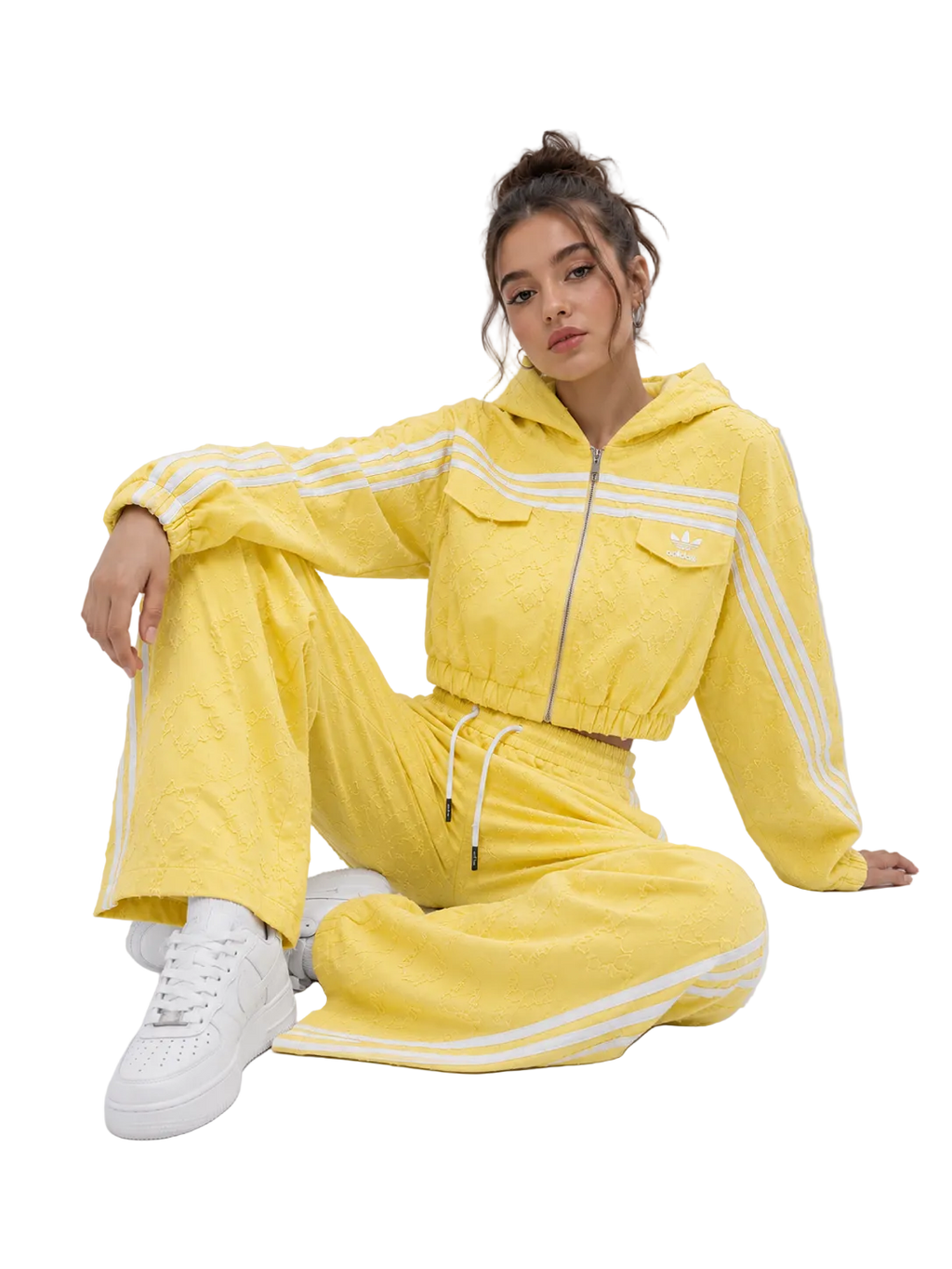 A model wearing a bright yellow textured tracksuit with white stripes, featuring a cropped zip-up hoodie and matching wide-leg pants, paired with white sneakers, set against a plain light gray studio background.