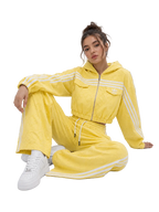 A model wearing a bright yellow textured tracksuit with white stripes, featuring a cropped zip-up hoodie and matching wide-leg pants, paired with white sneakers, set against a plain light gray studio background.