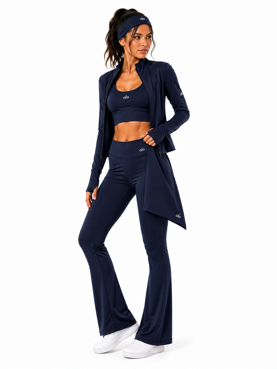 A woman modeling a matching navy blue activewear set consisting of a long-sleeve zip cardigan, sports bra, and high-waisted flared leggings, along with a matching headband and white sneakers, against a plain white background.