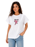 A woman modeling a white Balmain Paris graphic t-shirt with a pink flamingo print and black logo text, paired with light blue jeans against a plain white background.