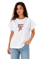 A woman modeling a white Balmain Paris graphic t-shirt with a pink flamingo print and black logo text, paired with light blue jeans against a plain white background.