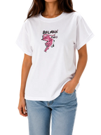 A woman wearing a white Balmain Paris graphic t-shirt with a pink flamingo print and black logo text, paired with light blue jeans, against a plain white background.