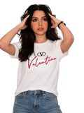 A woman wearing a white short-sleeve graphic t-shirt with a black Valentino logo and red script text, paired with blue jeans, posing against a light gray paneled wall.