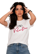 A woman wearing a white short-sleeve graphic t-shirt with a black Valentino logo and red script text, paired with blue jeans, posing against a light gray paneled wall.