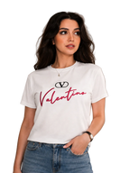 A woman wearing a white short-sleeve t-shirt with a red and black Valentino logo graphic on the front, paired with blue jeans, standing against a neutral beige paneled wall.